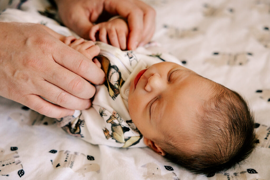 A newborn baby sleeps on a blanket with dad's hands holding it's hands