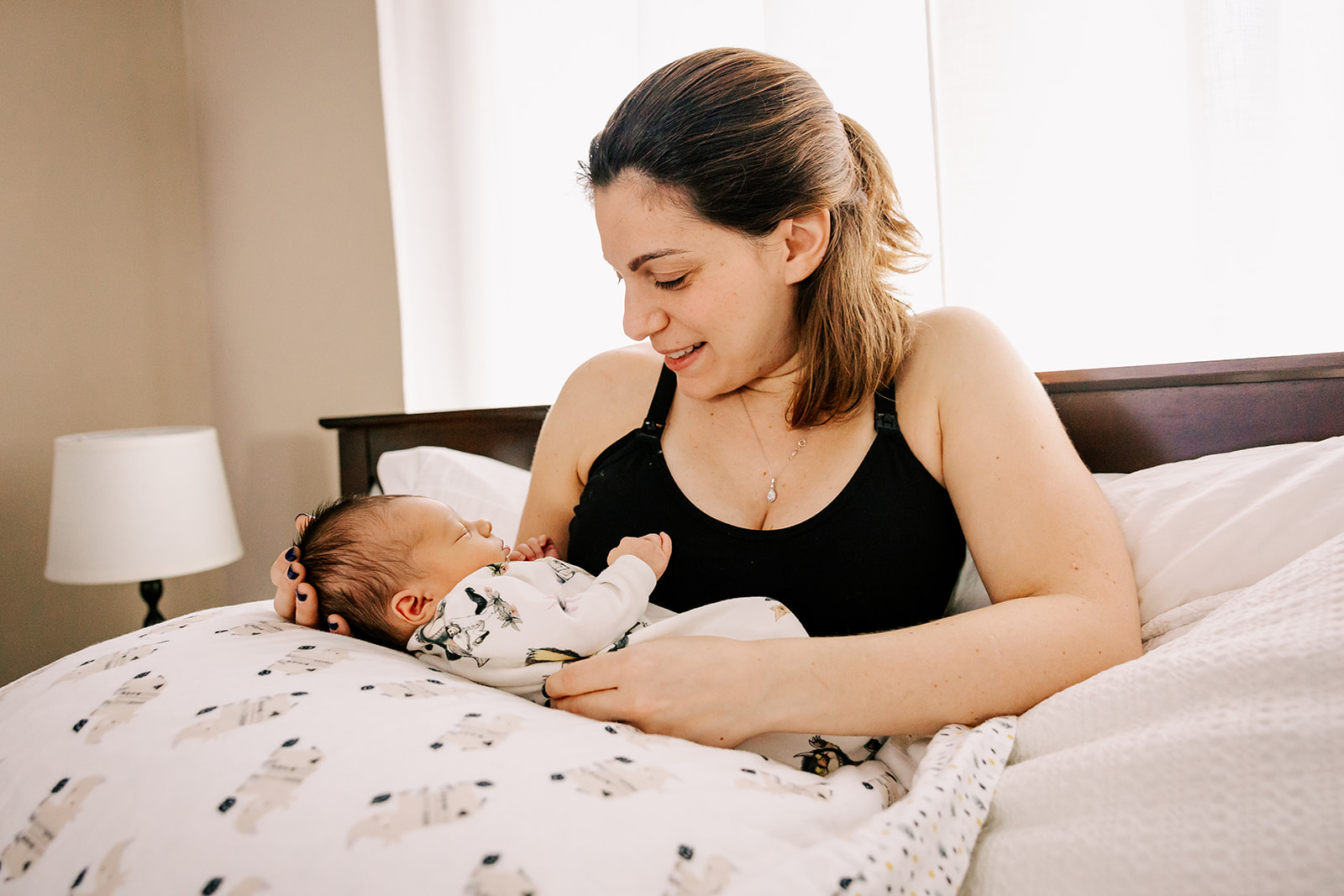 A happy mom lays in a bed holding her newborn in her lap smiling down at it after using a Midwife in Boston