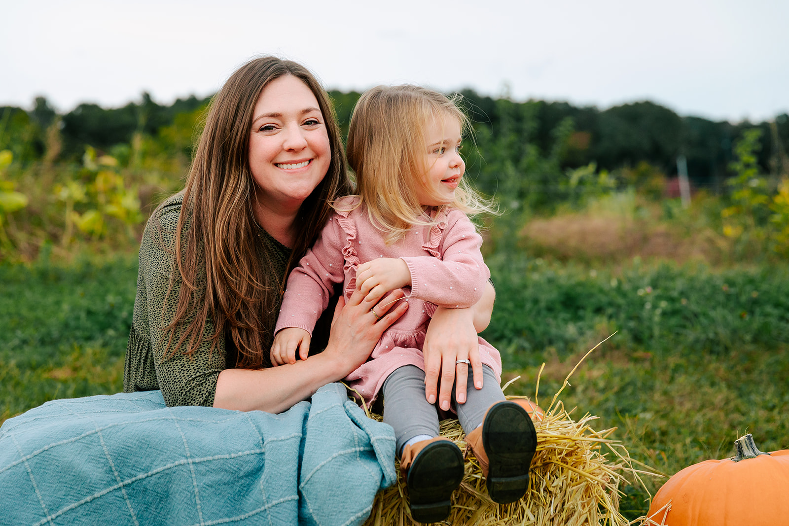 A happy mom smiles while sitting with her toddler daughter on a bail of hay in a park with a pumpkin before some mommy and me yoga in Boston