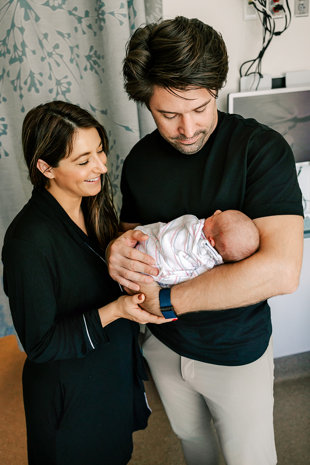 Happy new parents stand in a hospital room smiling down at their newborn baby in dad's arms