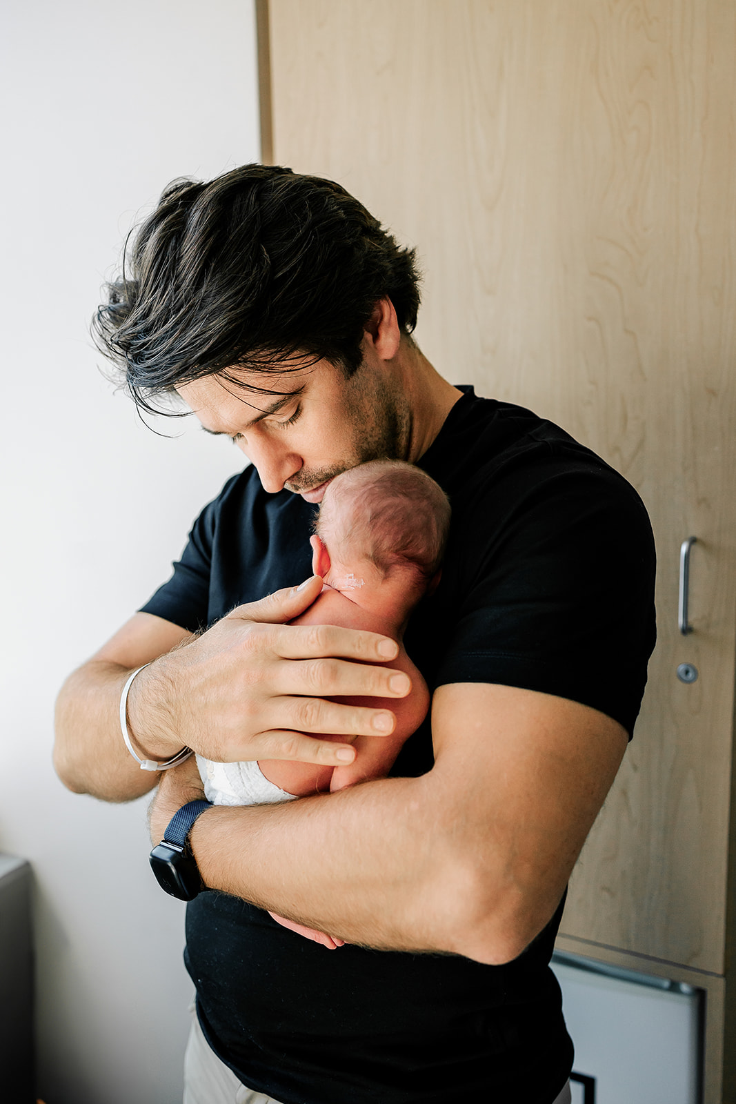 A new dad in a black shirt stands in the hospital cradling his newborn against his chest