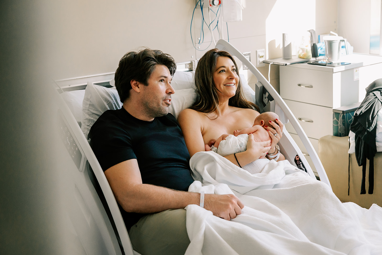 Happy new parents smile while laying in a hospital bed and breastfeeding their newborn after parenting classes in Boston