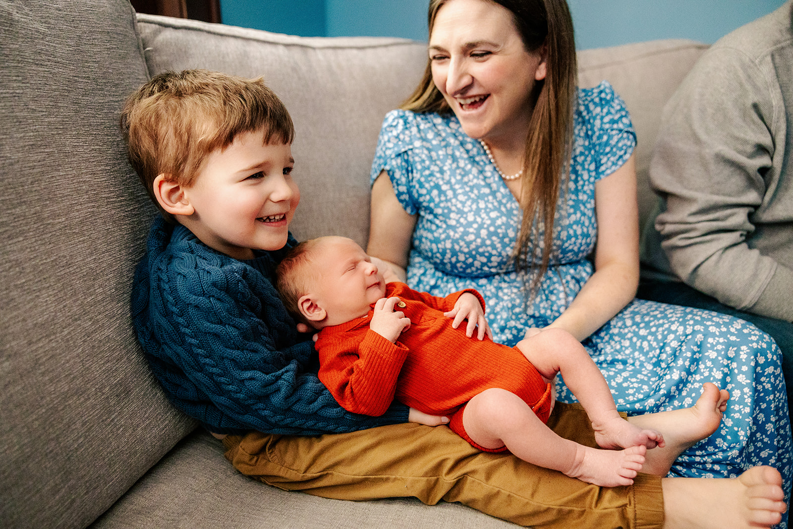 A toddler boy smiles while mom places his newborn sibling in his lap on a couch