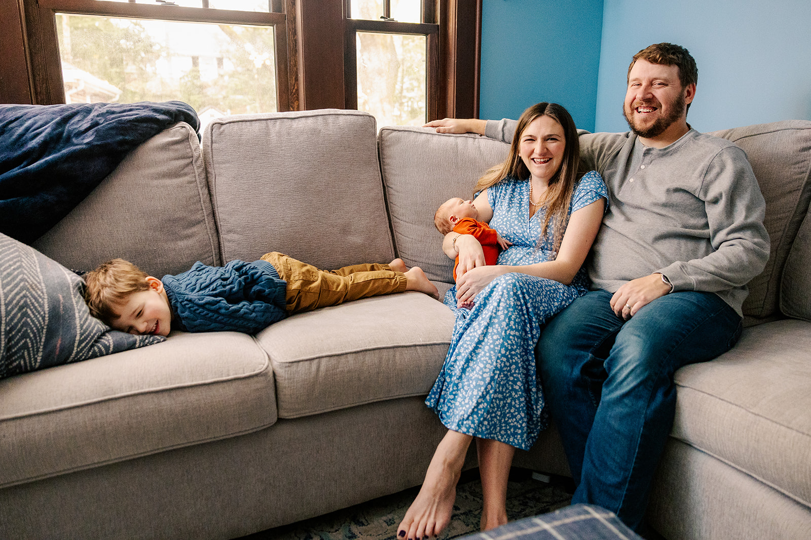 A mom and dad laugh while sitting on a couch holding a sleeping newborn as their toddler plays