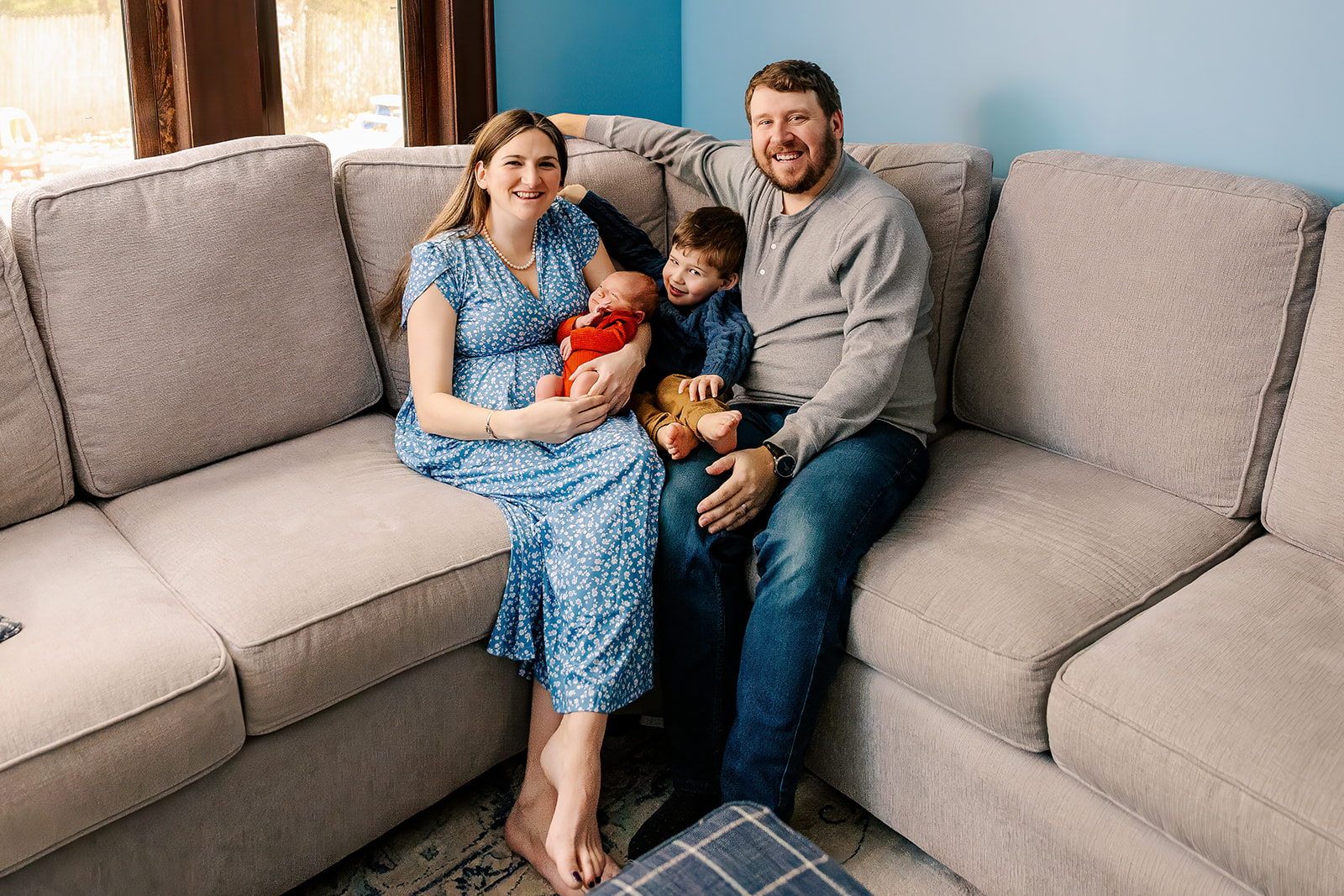 A happy family sits on a couch smiling after visiting a pediatric dentist in Boston with a newborn and toddler in mom and dad's lap