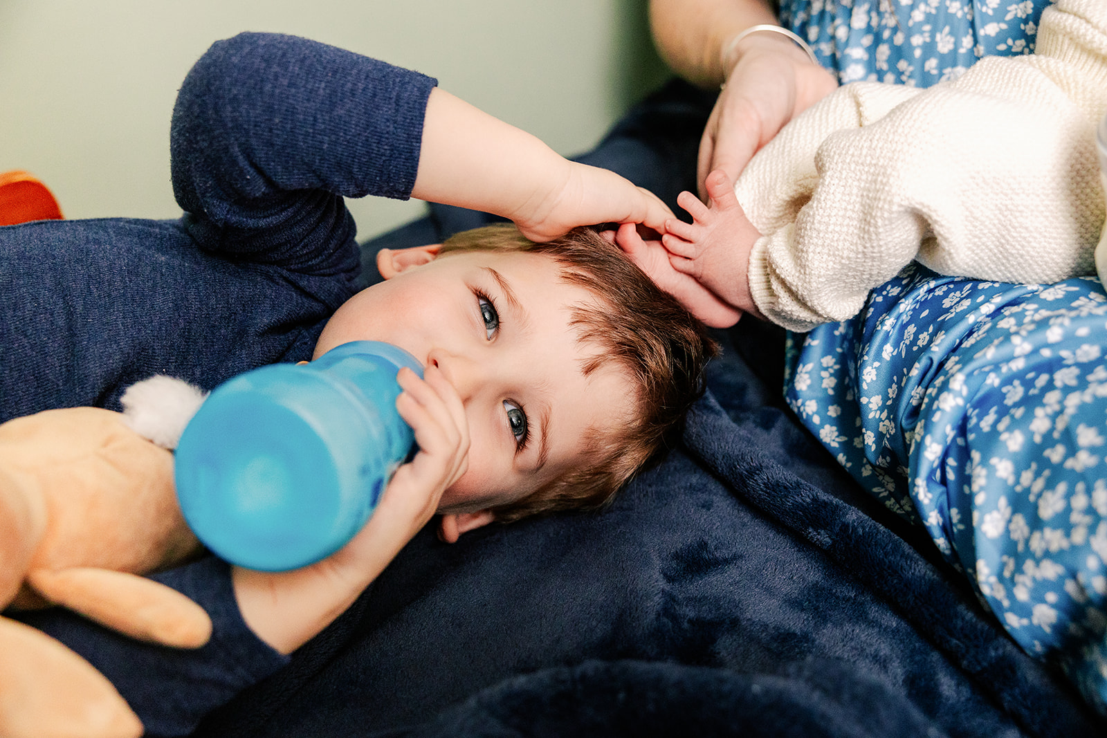 A toddler boy lays on a bed with his newborn sibling's feet on his head while drinking a bottle after visiting a pediatric dentist in Boston