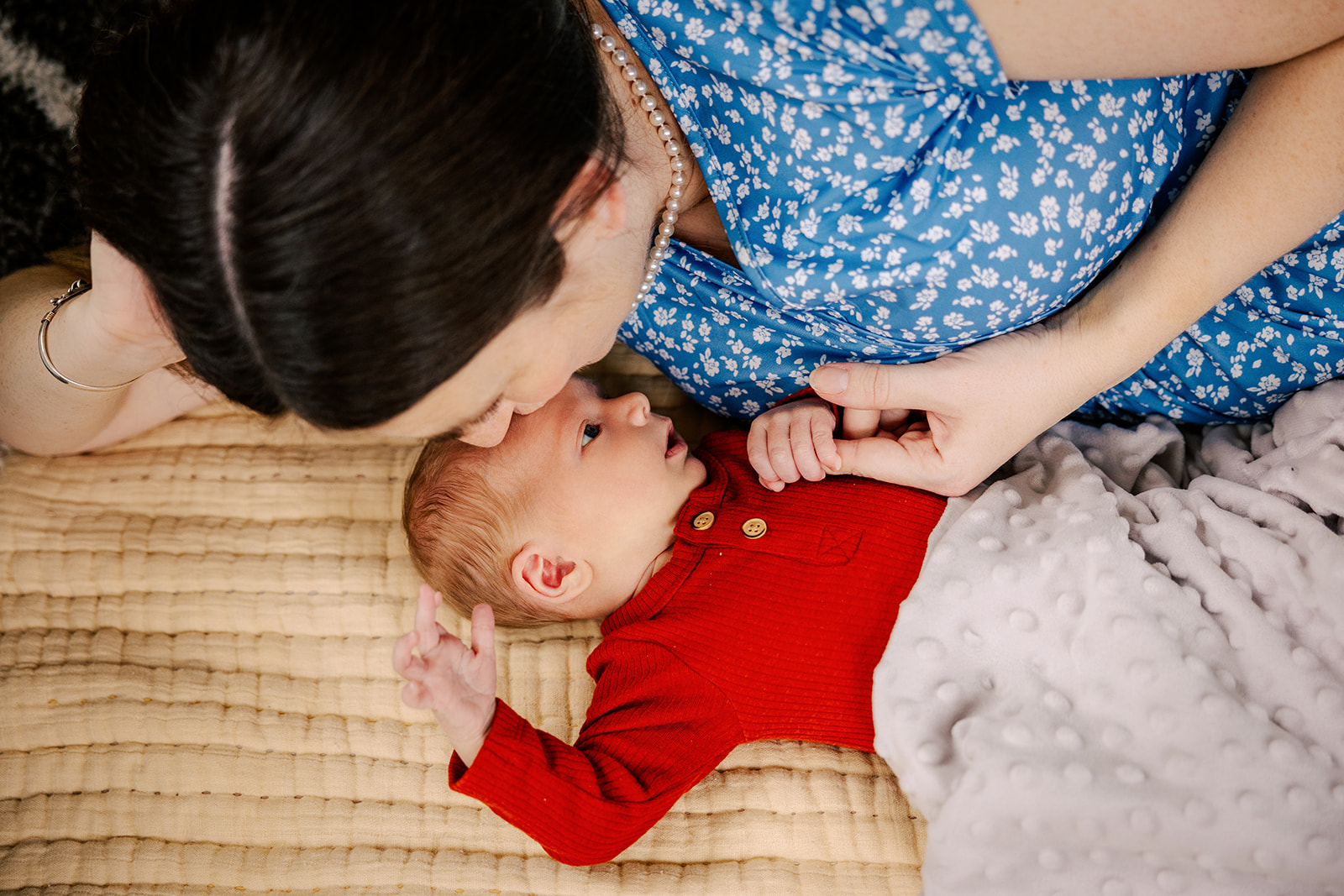 A newborn baby holds mom's finger while snuggling on a bed in a red onesie