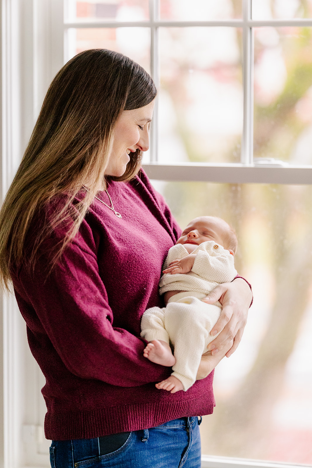 A new mom cradles her newborn in her arms while standing in a window in a purple sweater before some pelvic floor therapy in Boston