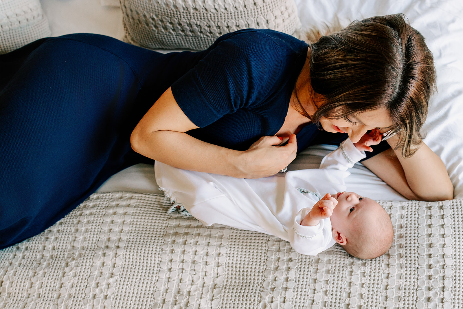 A new mom in a blue dress lays across a bed playing with her newborn baby in a white onesie