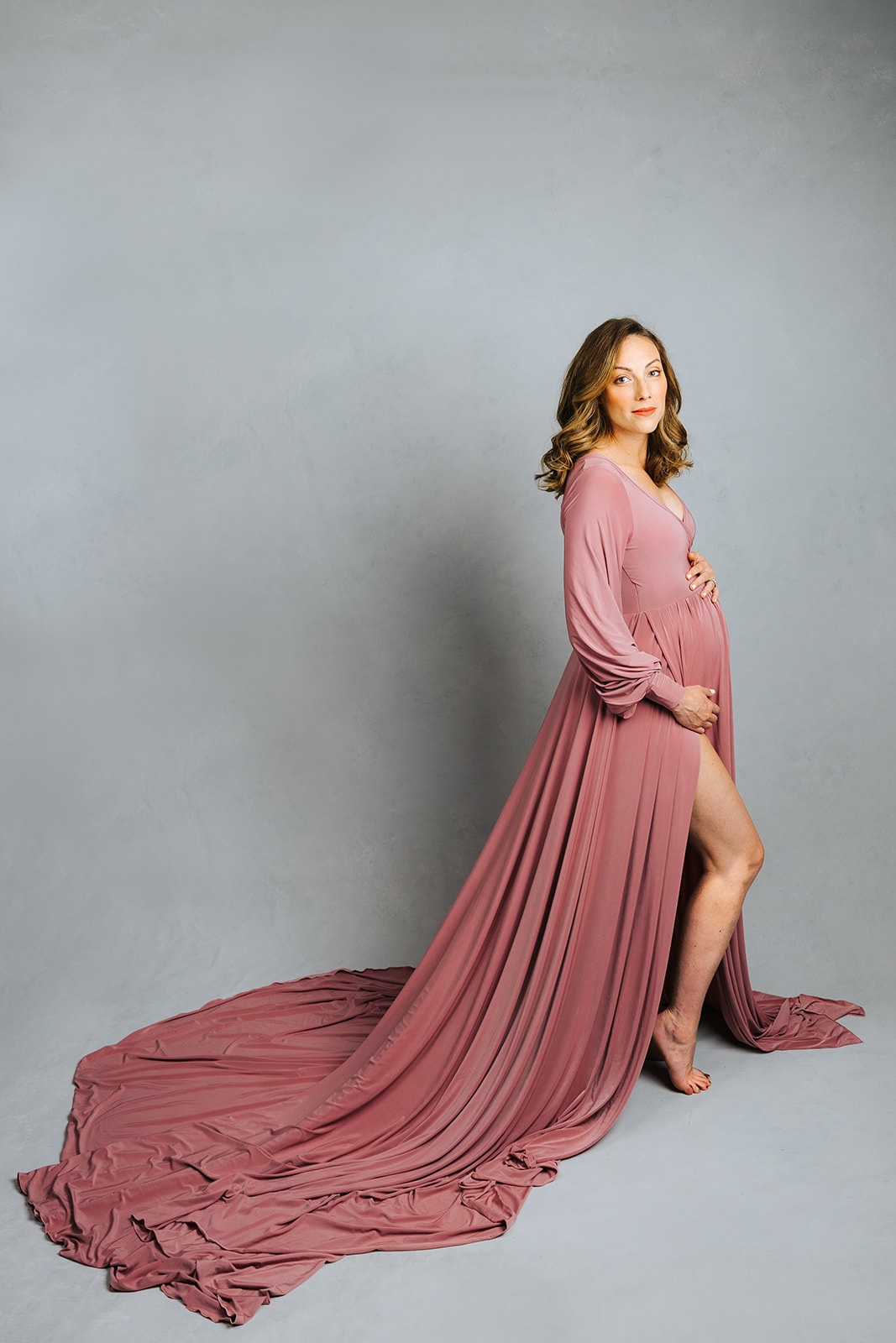 A happy pregnant woman stands in a studio with hands on her bump in a long pink maternity gown