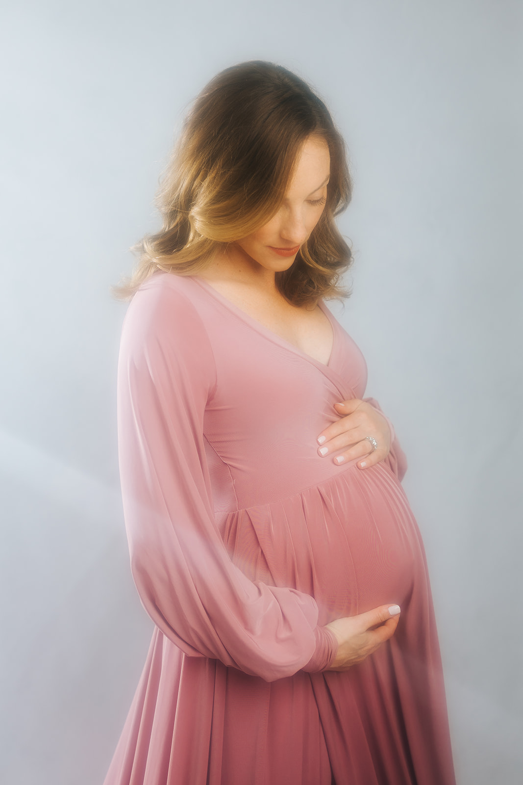 A mother to be holds her bump while looking down to it while standing in a studio after visiting a prenatal chiropractor in Boston