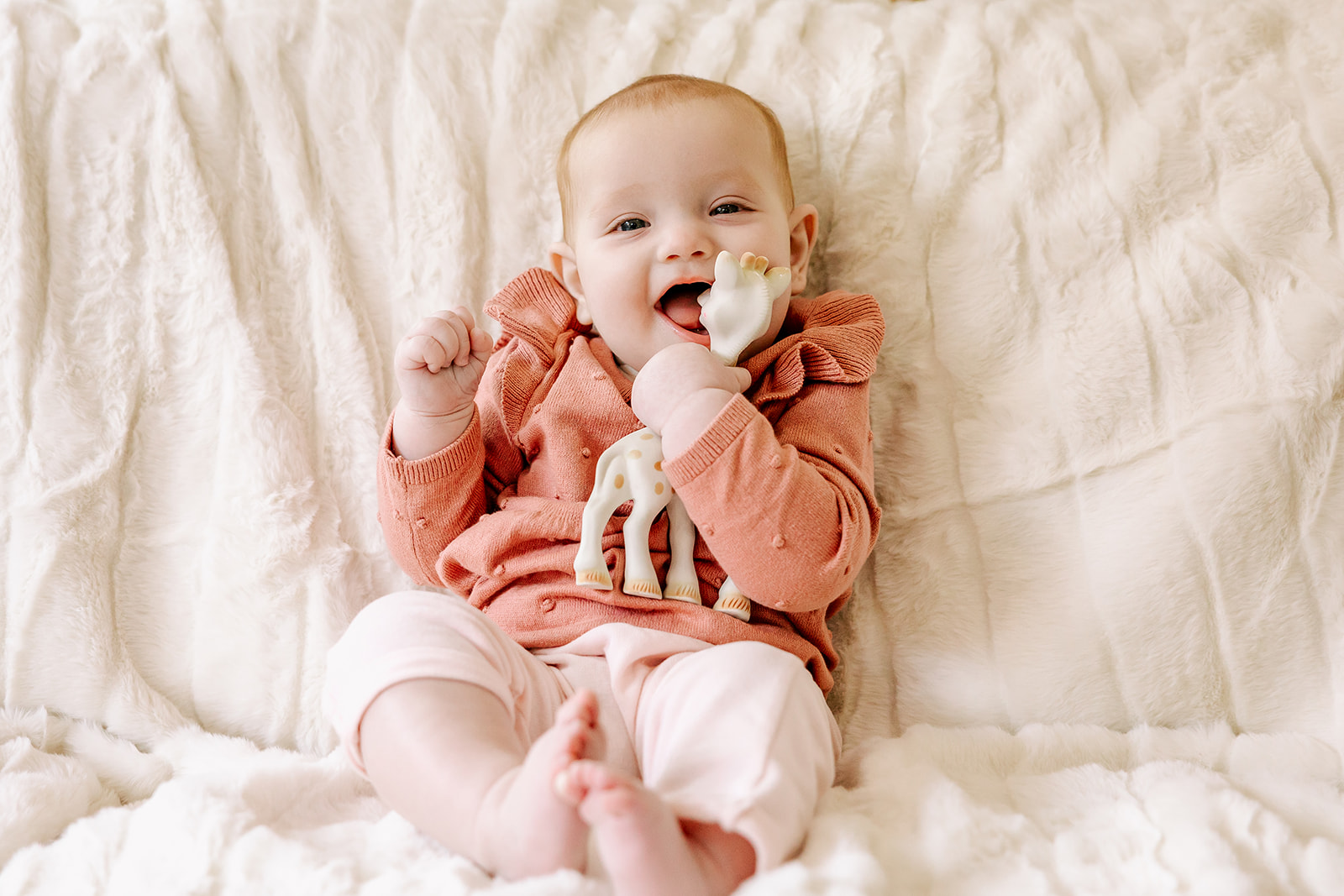 A happy baby girl in a pink sweater chews on a toy giraffe while lounging on a bed