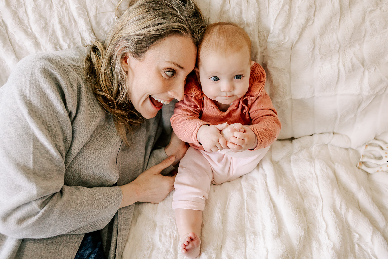 A happy mom plays with her baby in a grey sweater and pink sweater on a white bed with big smiles after some baby swim lessons in Boston