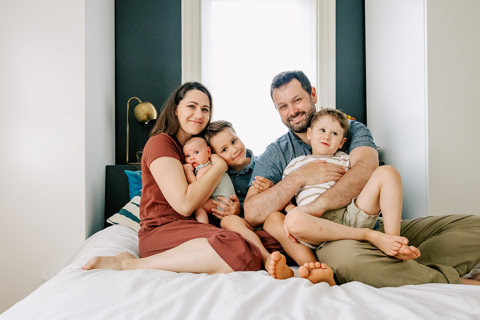 A mom and dad sit on a bed snuggling their baby and two toddler sons with big smiles