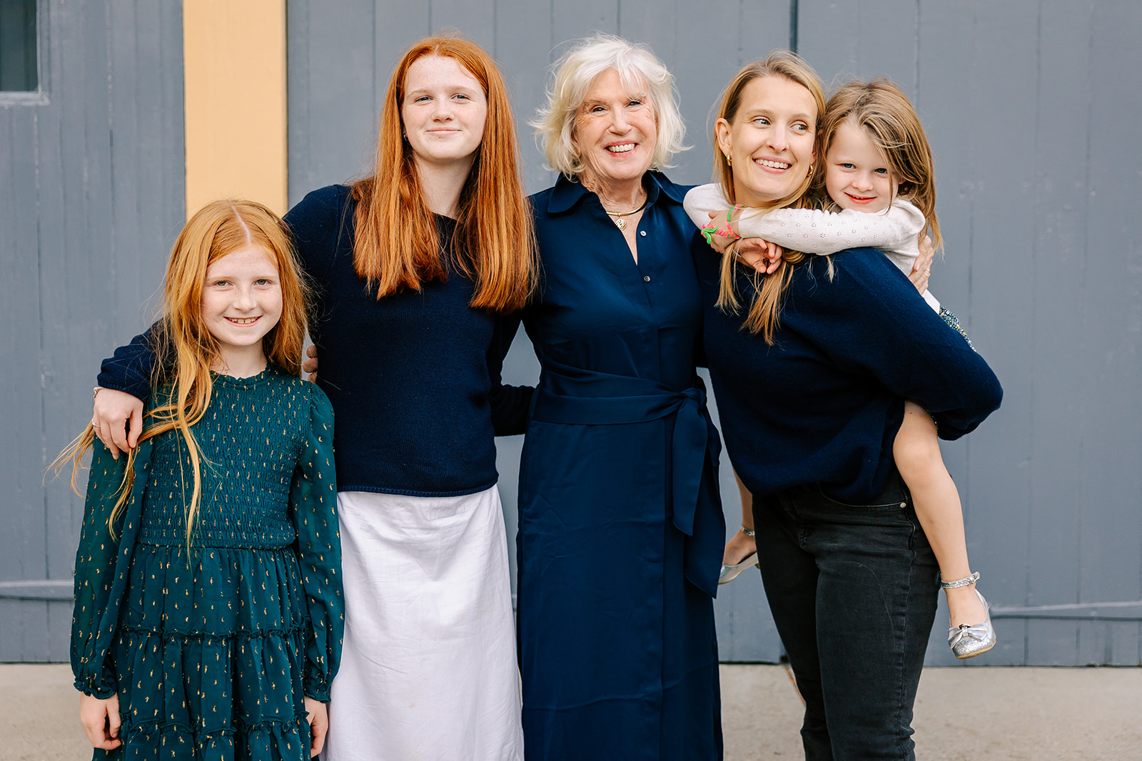 A smiling grandma stands in a blue dress with her daughter and three grand daughters