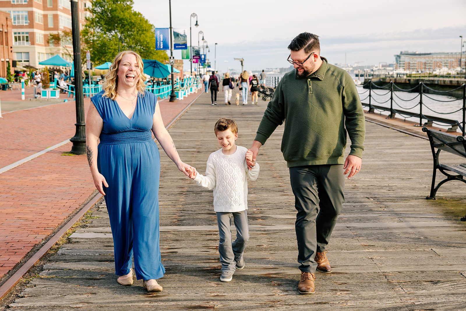 Happy parents laugh while walking the wharf boardwalk at sunset holding hands with their child after some kids birthday parties in Boston