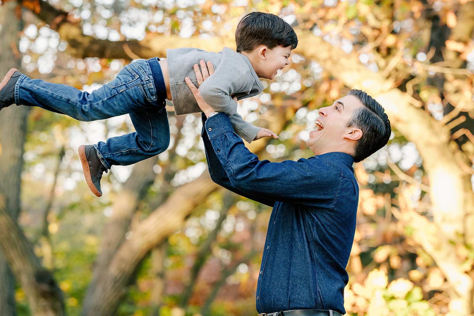 A dad laughs while lifting his giggling toddler son in the air in a park at sunset