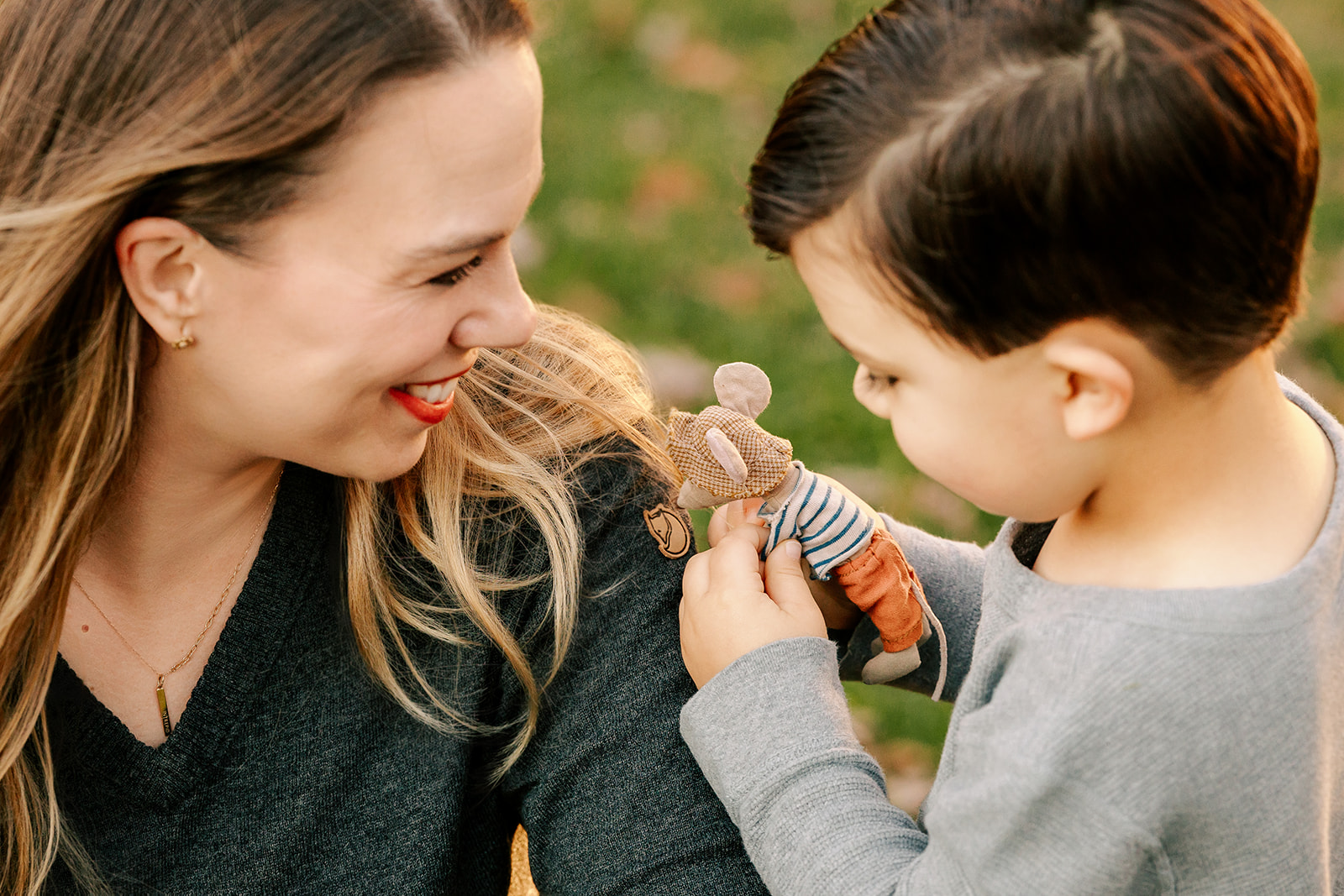 A young boy plays with a tiny stuffed mouse on mom's shoulder in a park