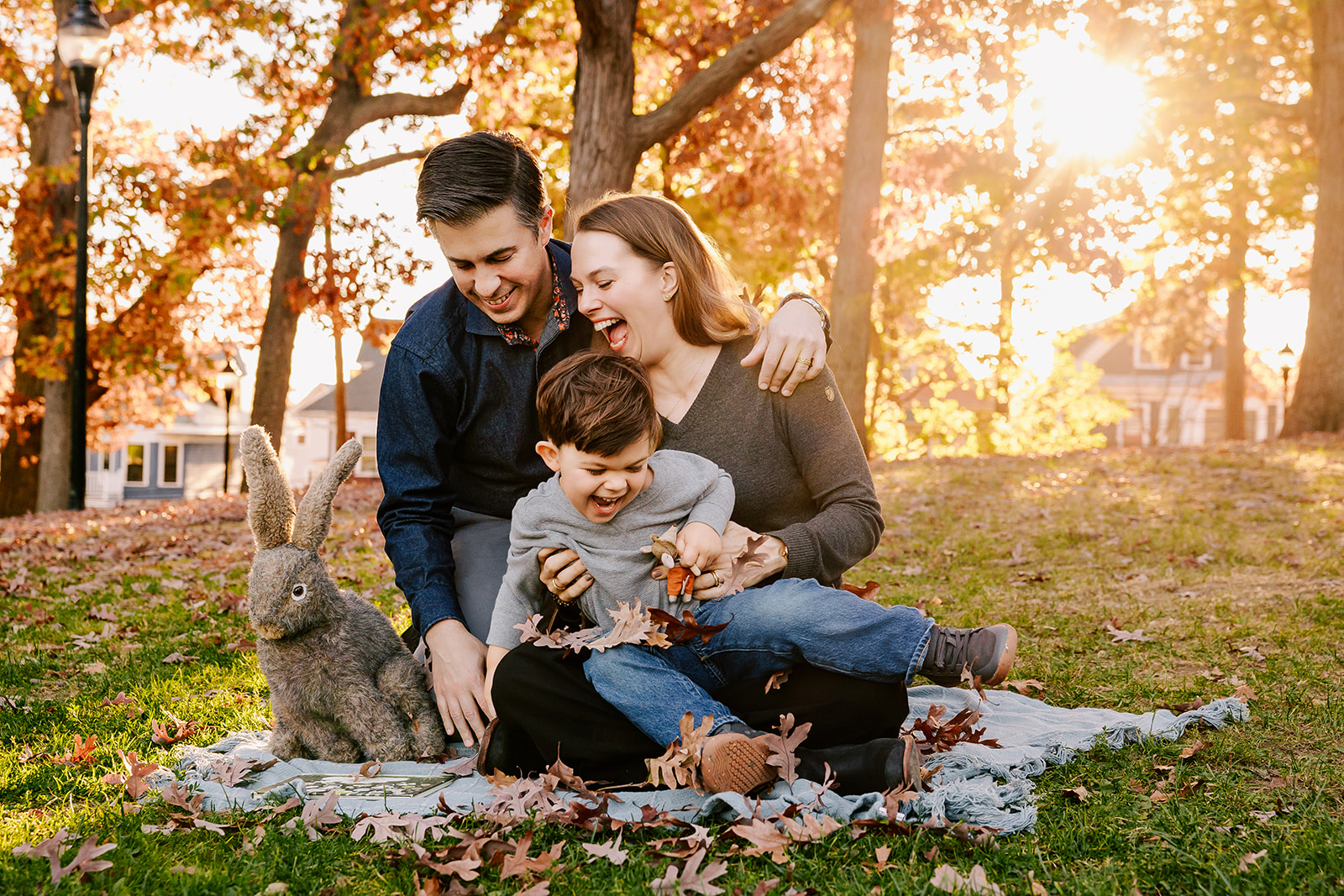 A happy mom and dad laugh while playing with their giggling toddler son in mom's lap in a park at sunset after some mommy and me classes in Boston