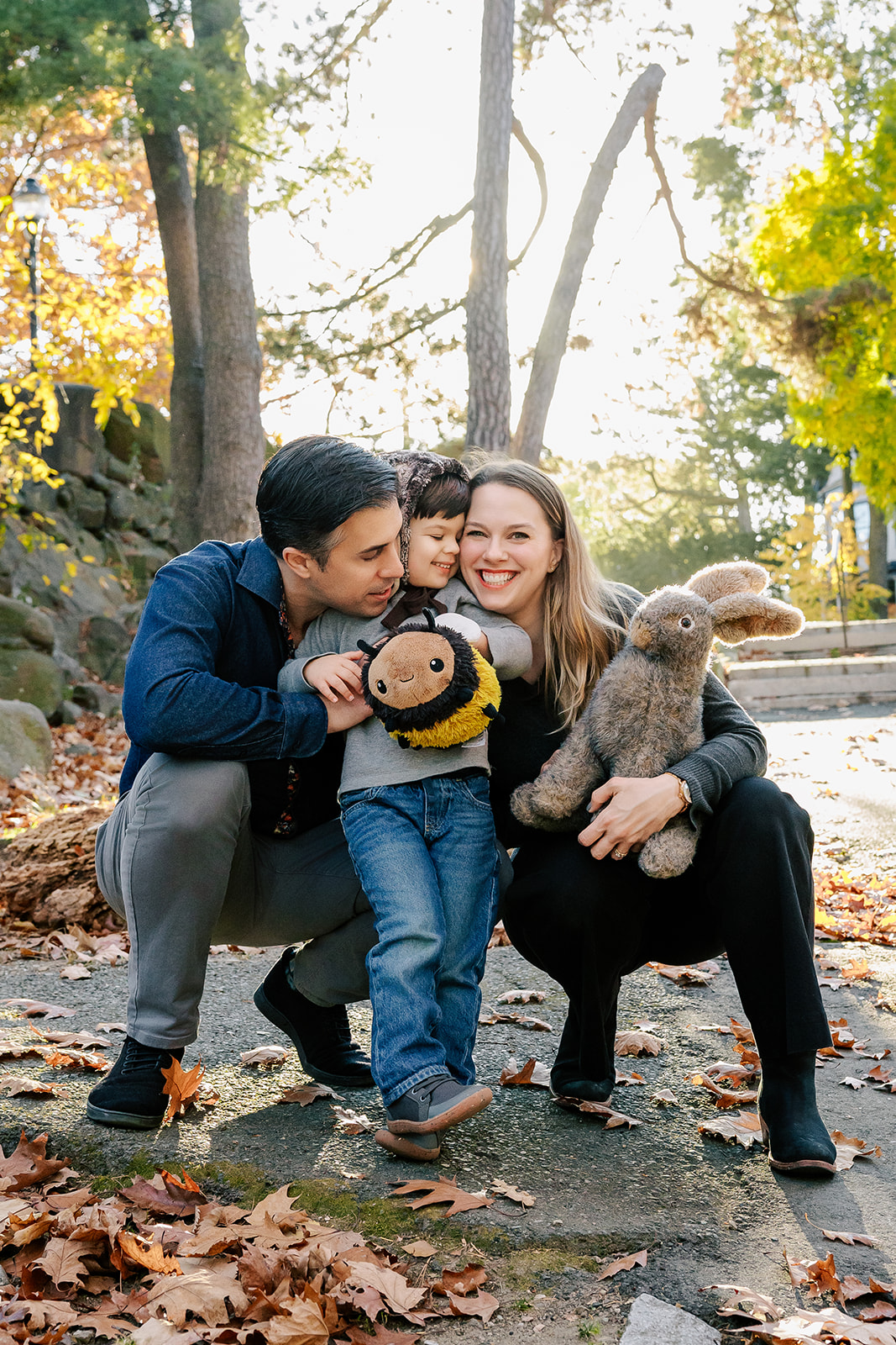 Happy mom and dad squat to hug and snuggle their toddler son in a park with stuffed animals after some mommy and me classes in Boston