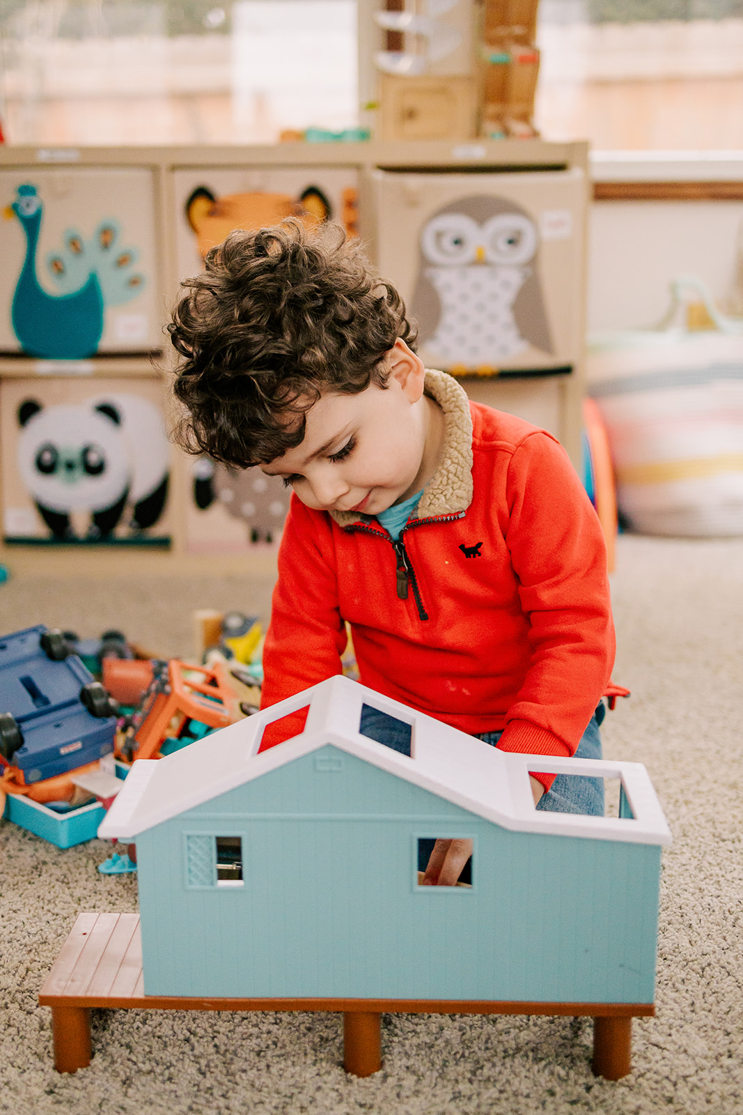 A young boy plays with a toy house in his home in a red sweater and a smile