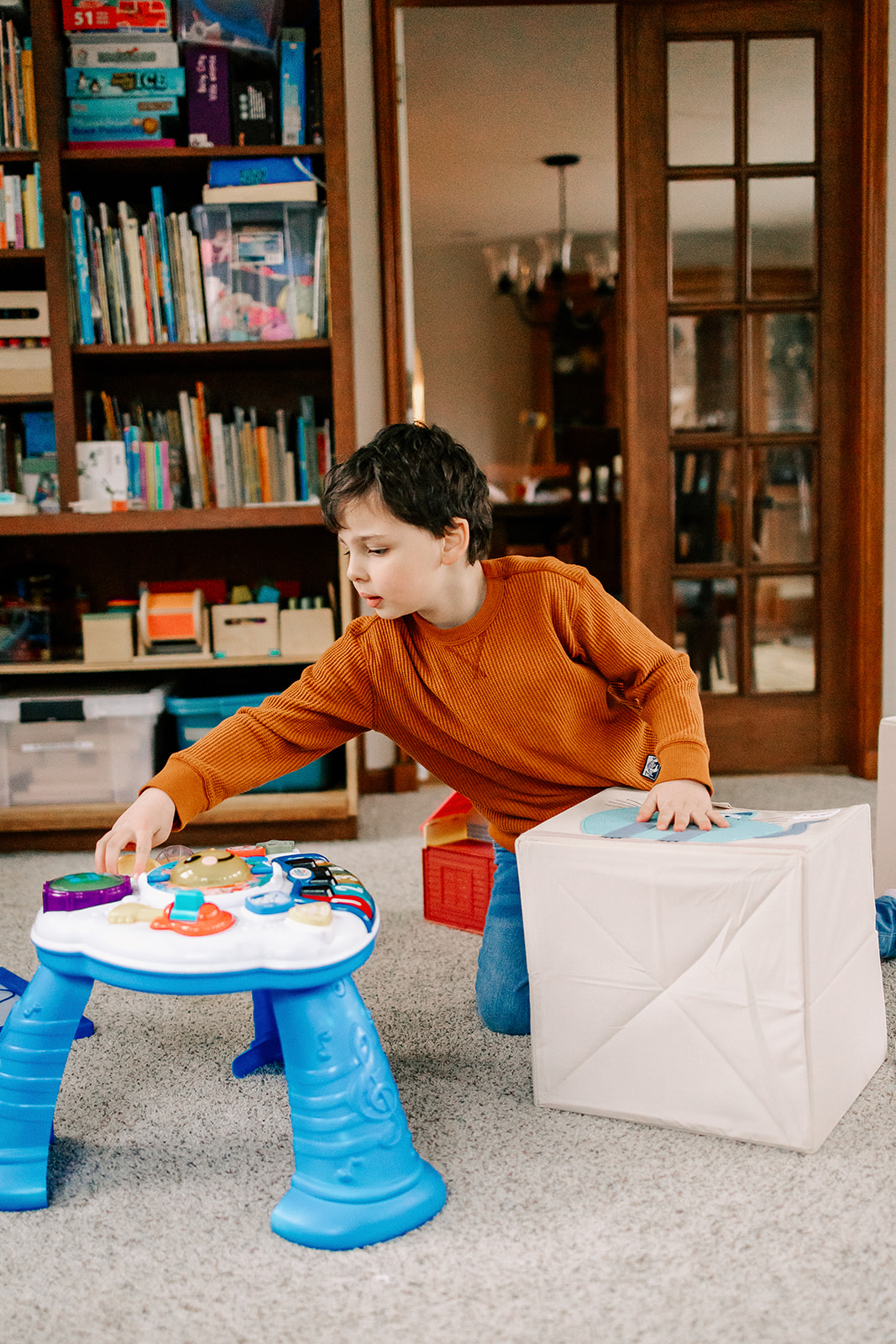 A young boy in an orange shirt and jeans plays with toys in a living room with a padded box and musical toy after visiting Montessori Schools in Boston