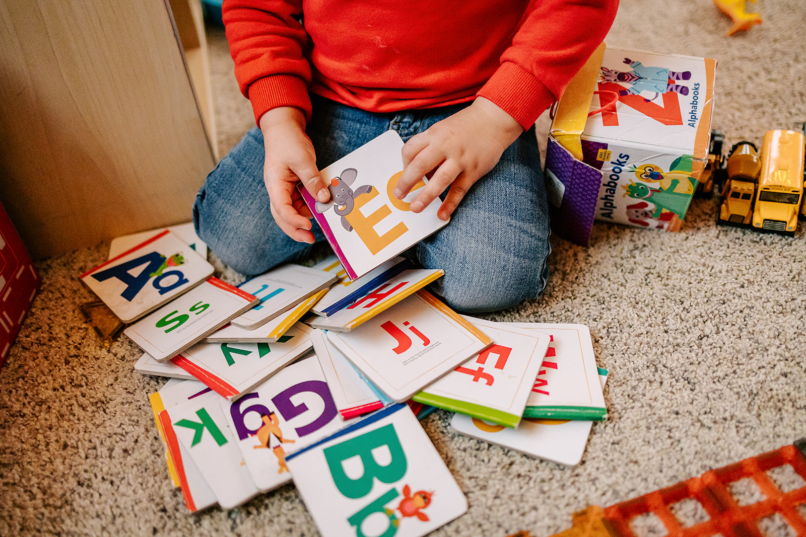 A toddler in a red sweater and jeans plays with a stack of letter books after visiting Montessori Schools in Boston