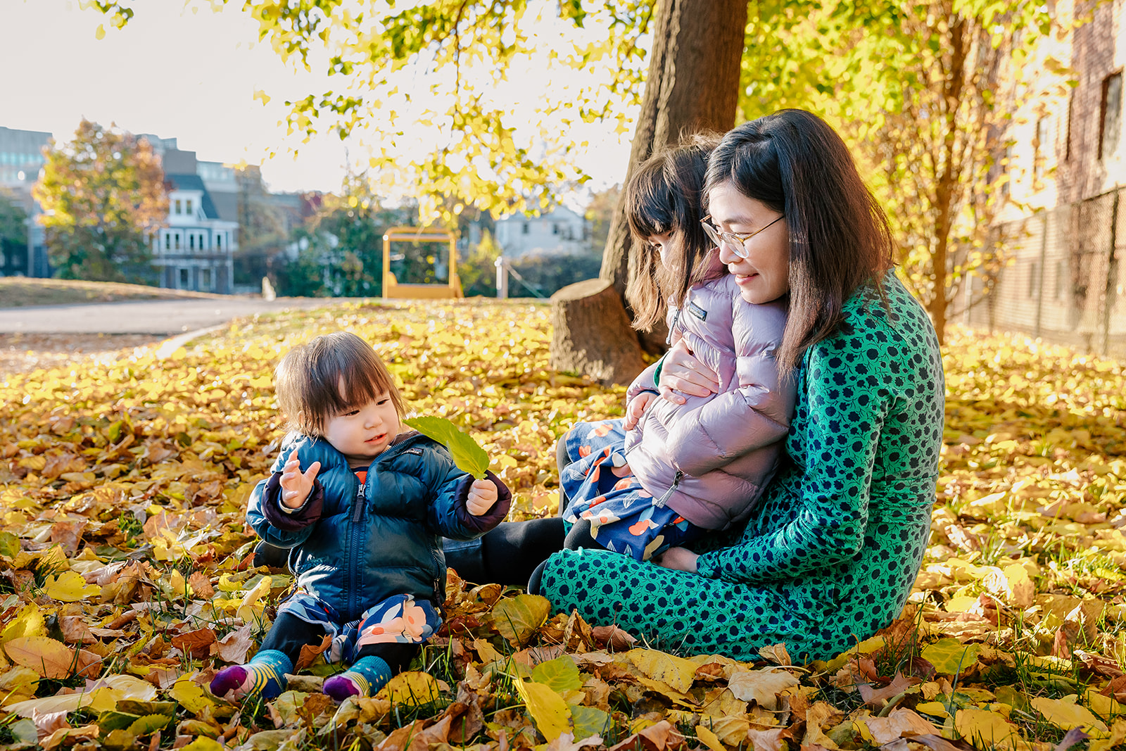 A happy mom sits in leaves in a park with her toddler daughters exploring the fallen leaves