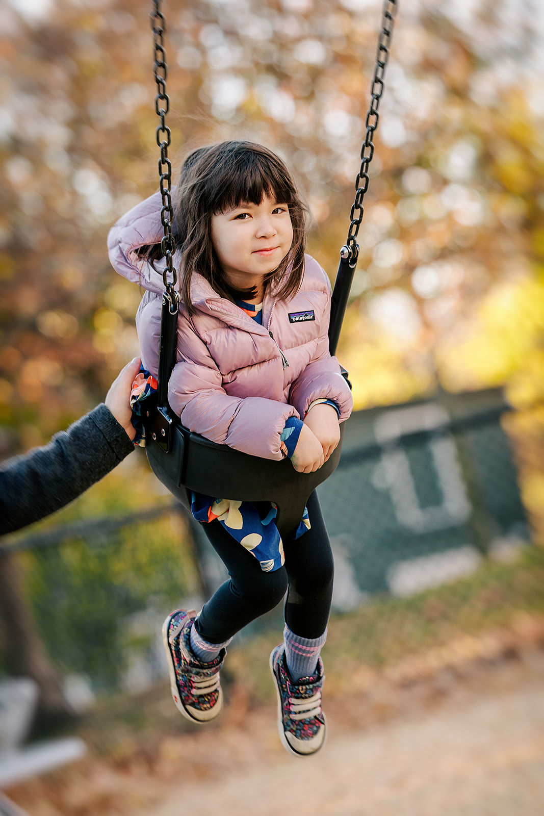 A young girl sits in a moving swing in a pink jacket with dad's help