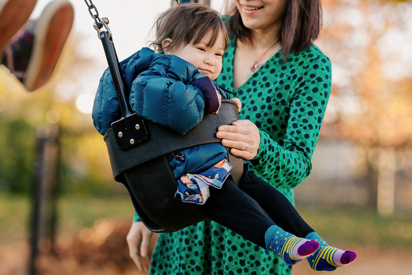 A happy toddler in a big jacket sits in a swing being pushed by mom at one of the Playgrounds in Boston