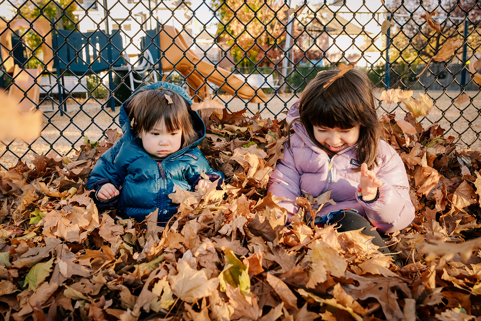 Toddler sisters in jackets play and laugh while sitting in a pile of leaves at one of the Playgrounds in Boston