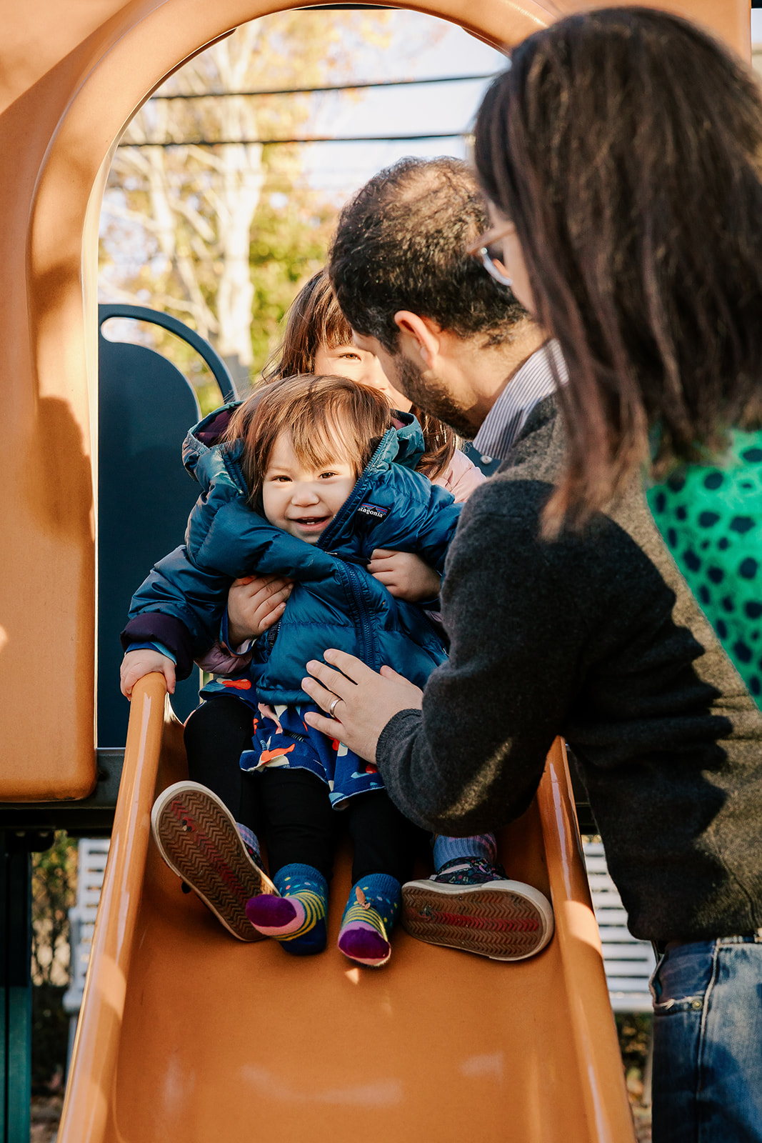 A toddler girl sits on a slide with her baby sister in her lap with mom and dad helping at one of the Playgrounds in Boston