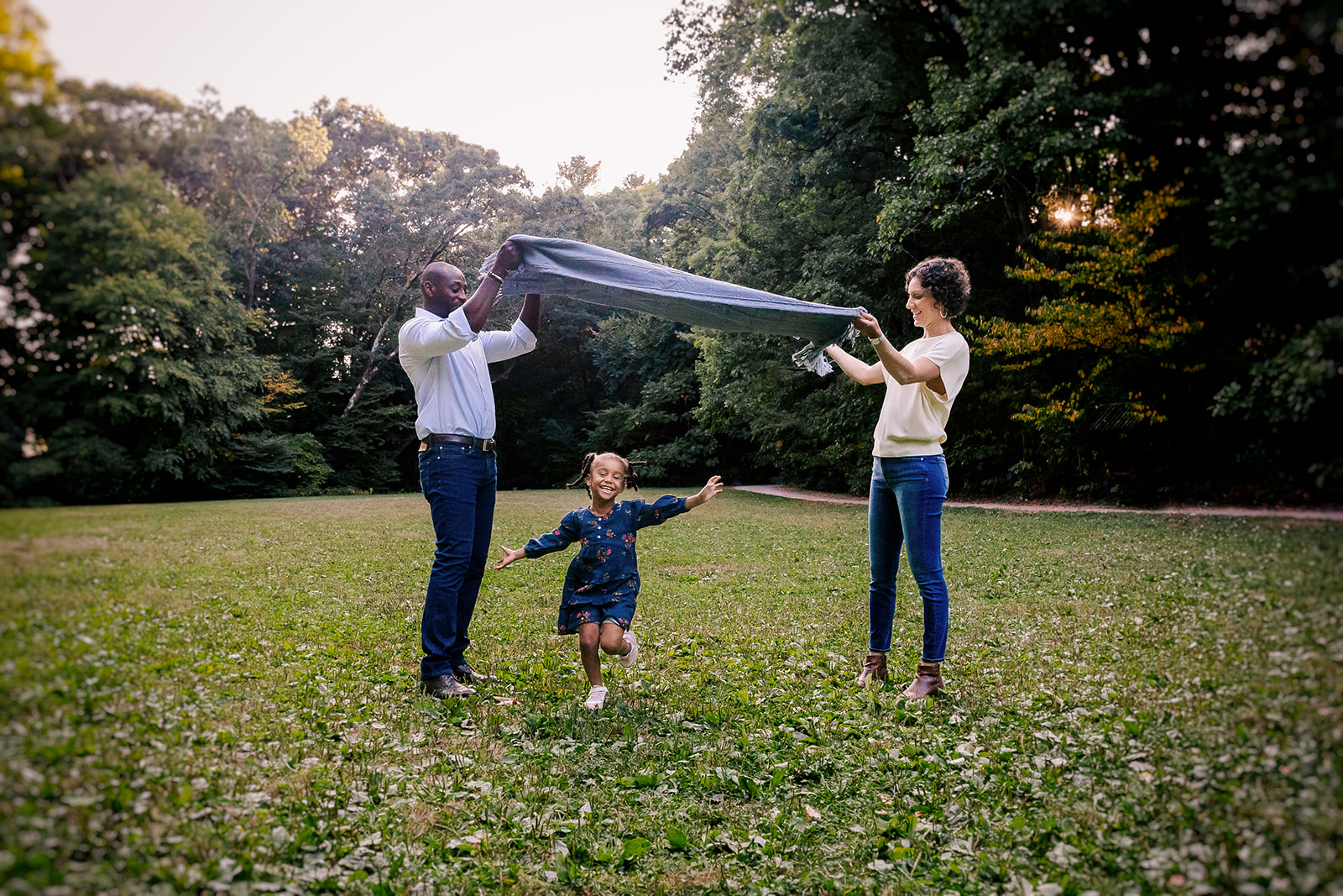 A toddler in a blue dress runs under a blanket held by mom and dad in a park lawn at sunset