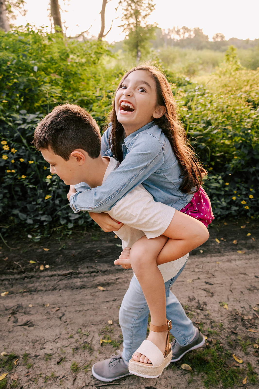 A young boy gives his younger sister a piggy back ride in a park trail at sunset