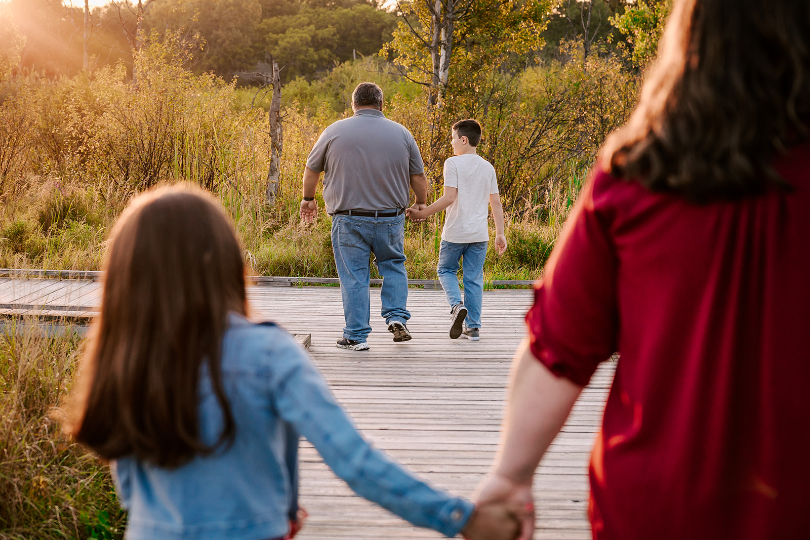 A mom and dad walk holding hands with their son and daughter on a boardwalk at sunset