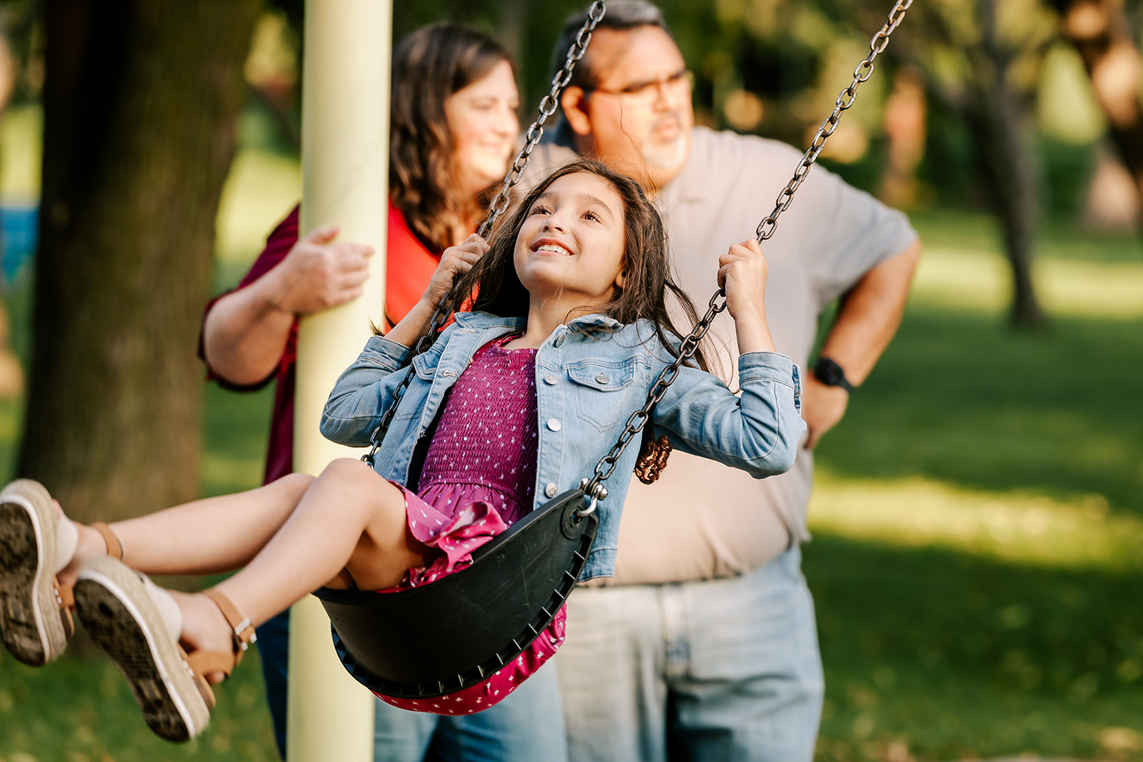 A young girl in a pink dress swings on a swingset in front of mom and dad after some summer camps in Boston