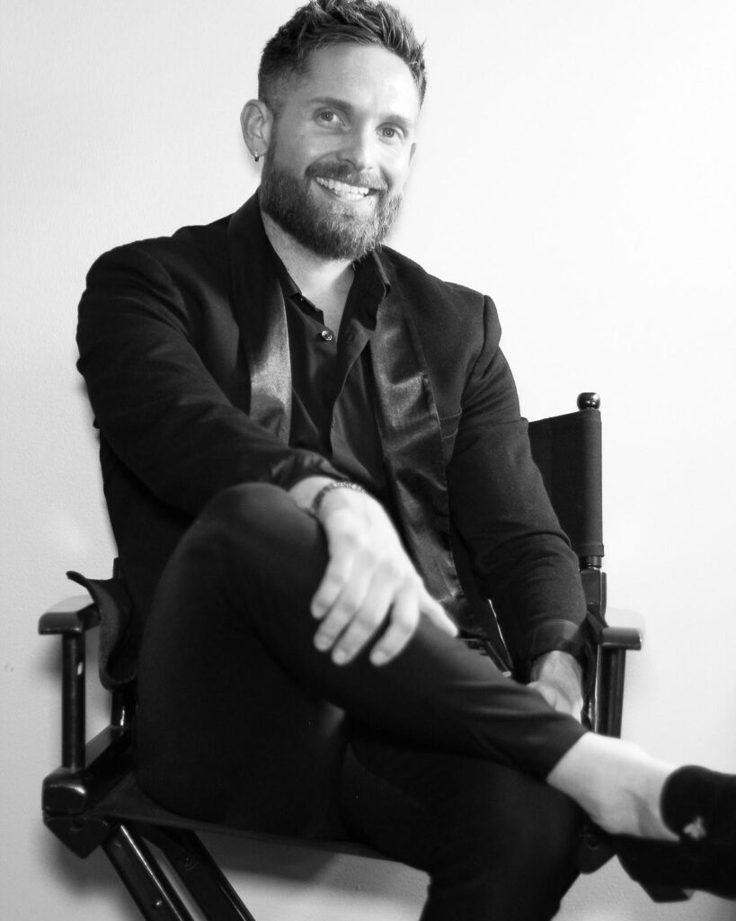 Black and white photo of a smiling bearded young man sitting in a director's chair.