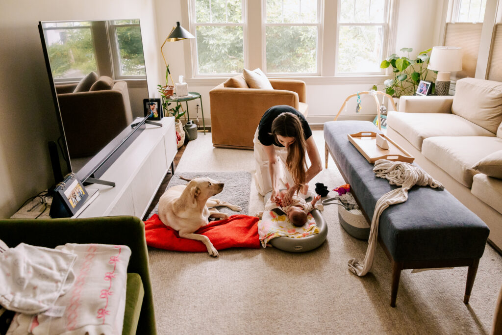 A golden lab looks up with concern at his adult owner, who is leaning over a changing pad with her week old baby.