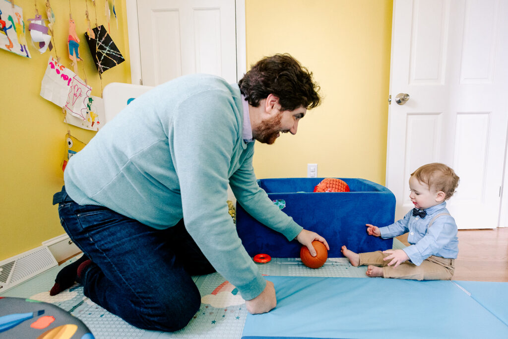 A happy dad in a blue sweater plays with his toddler son with balls after visiting doulas in Cambridge, MA