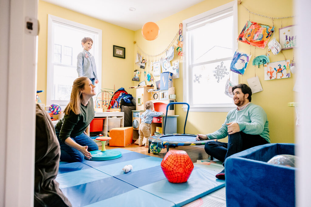 A mom and dad play with a balloon in a play room with their two toddler sons in blue shirts after visiting doulas in Cambridge, MA