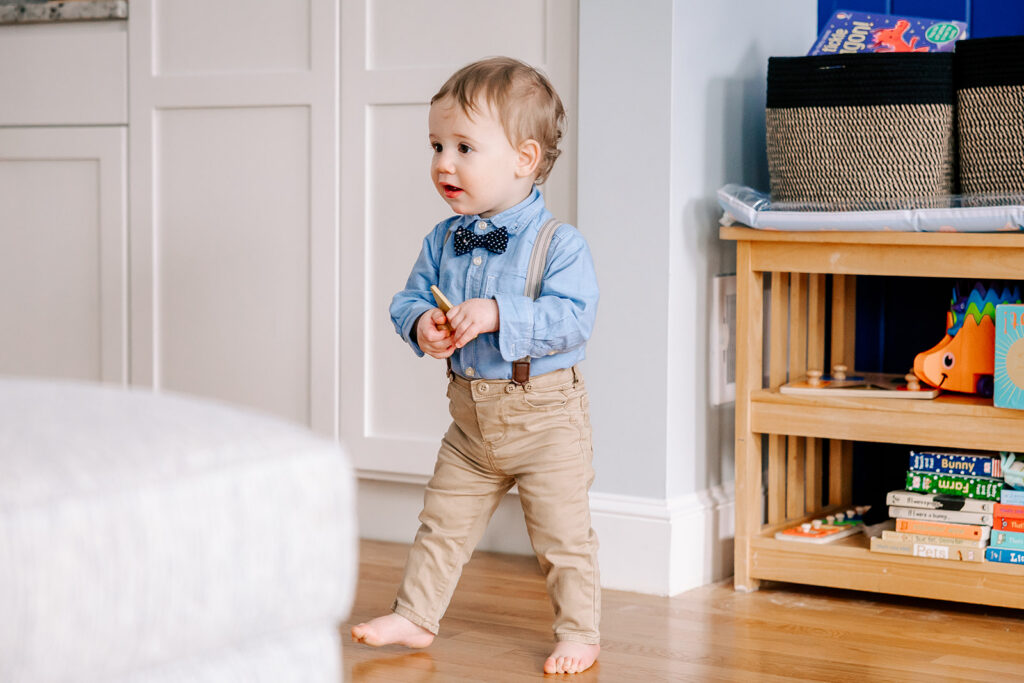 A toddler boy in a blue shirt, bowtie and suspenders walks with a wooden toy after visiting daycares in Cambridge, MA