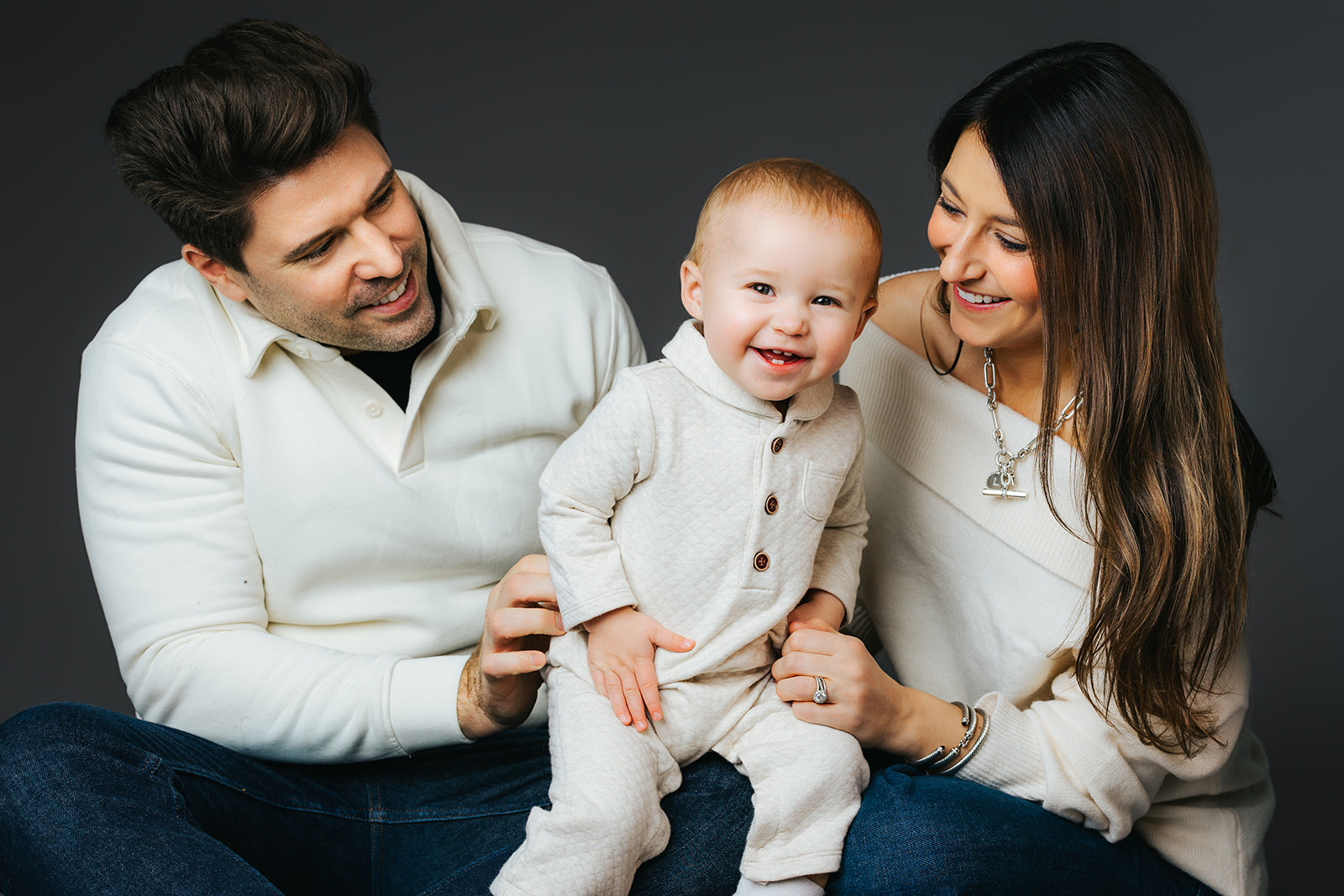 A mom and dad in white sweaters laugh while playing with their happy toddler son between them in a studio
