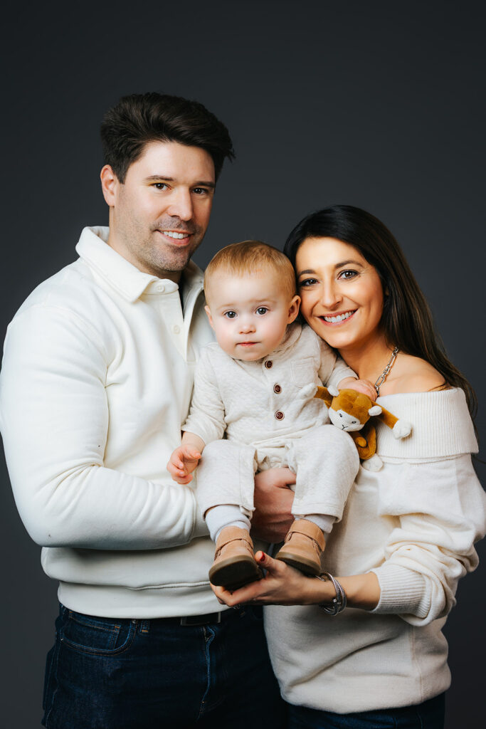 Smiling mom and dad stand in a studio in whtie sweaters matching their toddler son they are holding