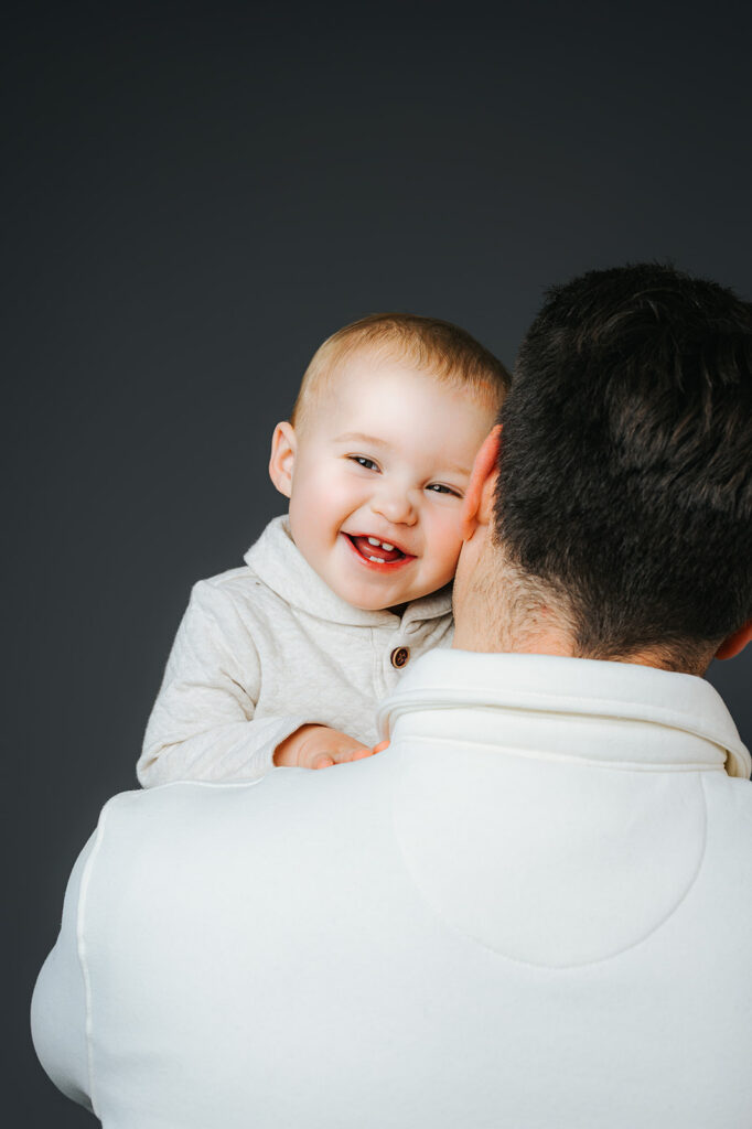 A giggling baby boy in a white sweater in dad's arms looking over his shoulder in a studio after finding a pediatric dentist in Cambridge, MA