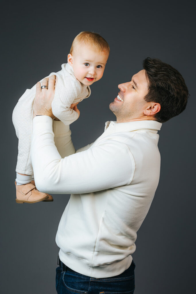 A happy dad in a white sweater lifts his giggling baby son in white in a studio after visiting a pediatric dentist in Cambridge, MA