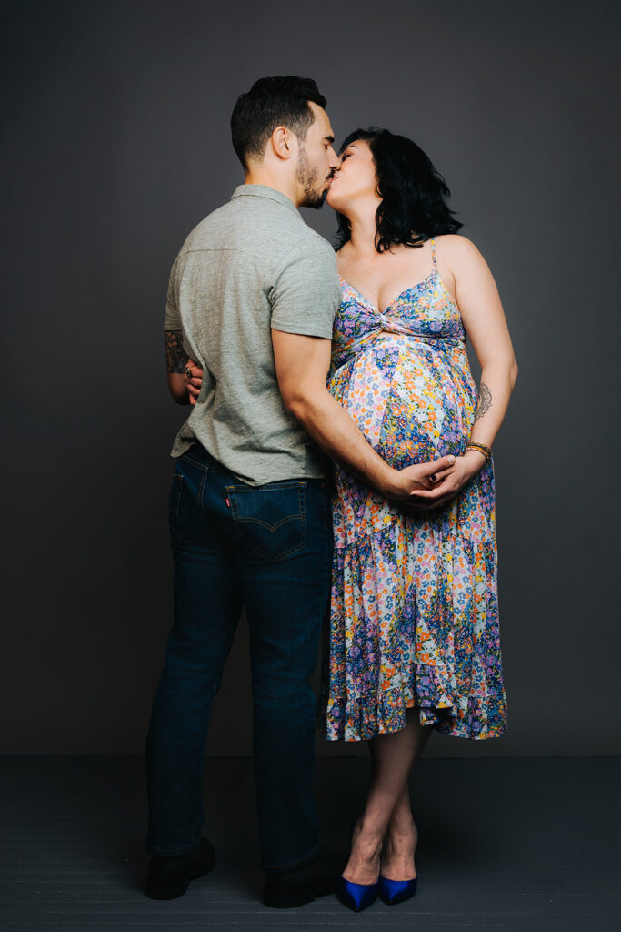 A couple kiss while holding hands under the bump while standing in a studio in bright colors
