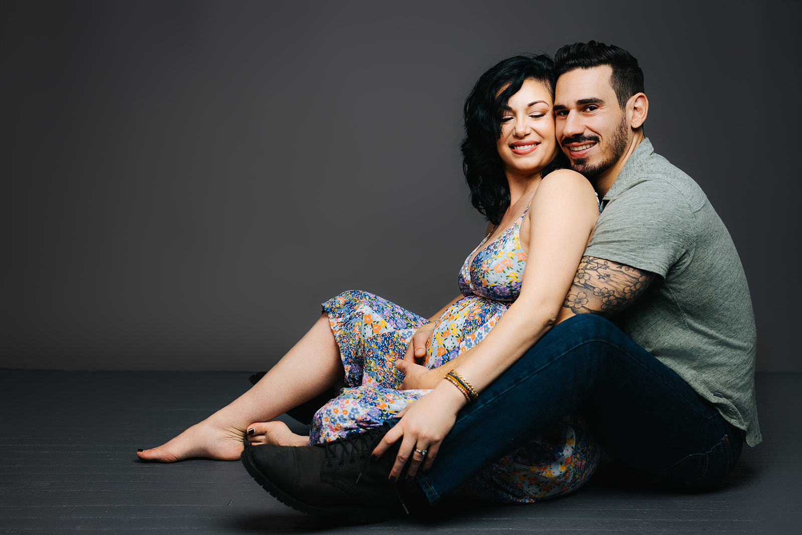 A pregnant woman sits in the lap of her husband on the floor in a bright flower print maternity dress