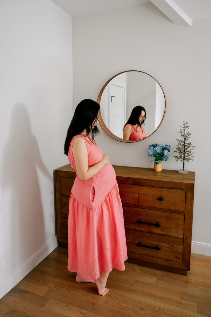 A soon to be mom stands in front of a dresser and mirror while looking down and holding her pregnant bump.