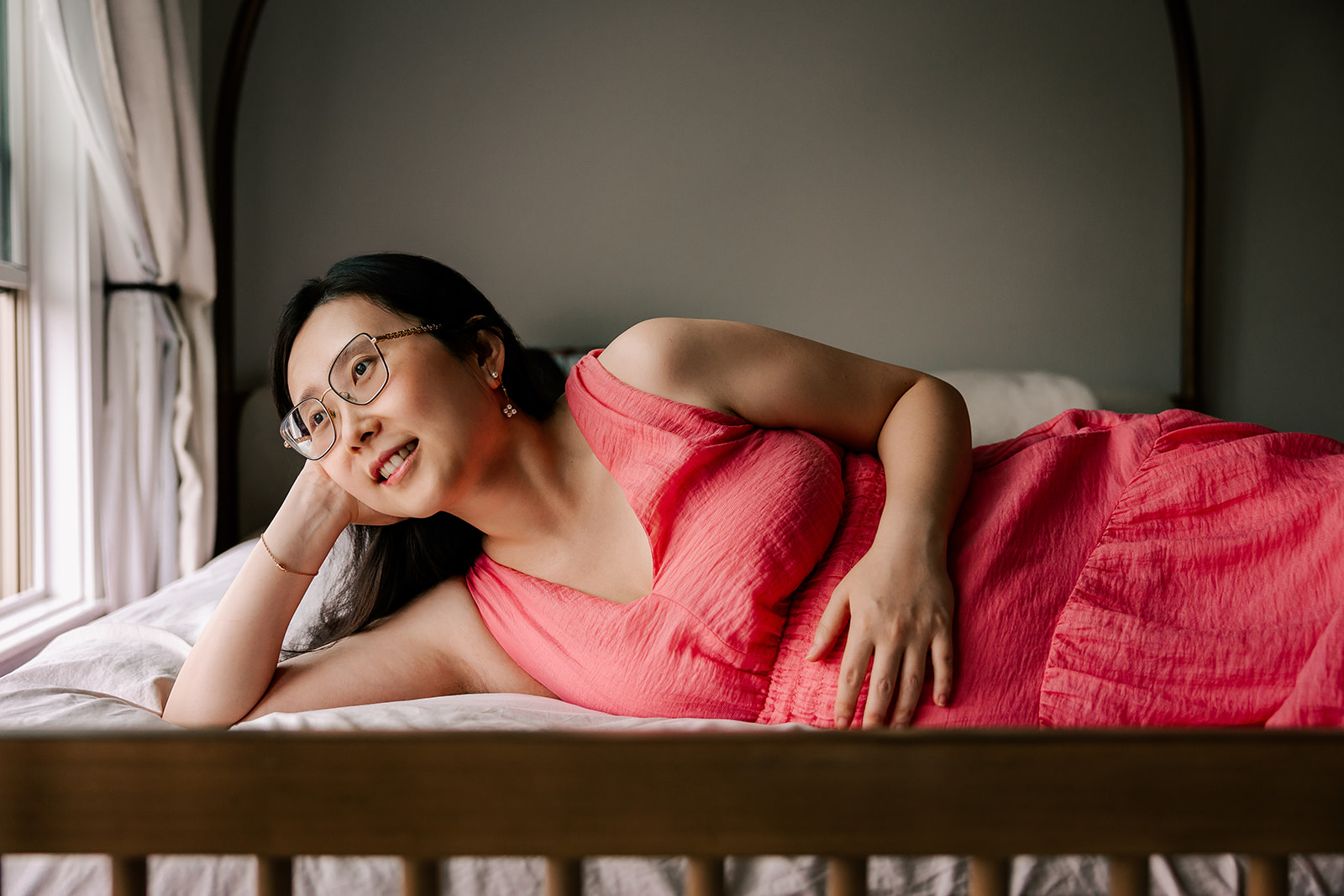 A pregnant mother in a pink dress lays on a bed looking out a window, with her hand on her bump.