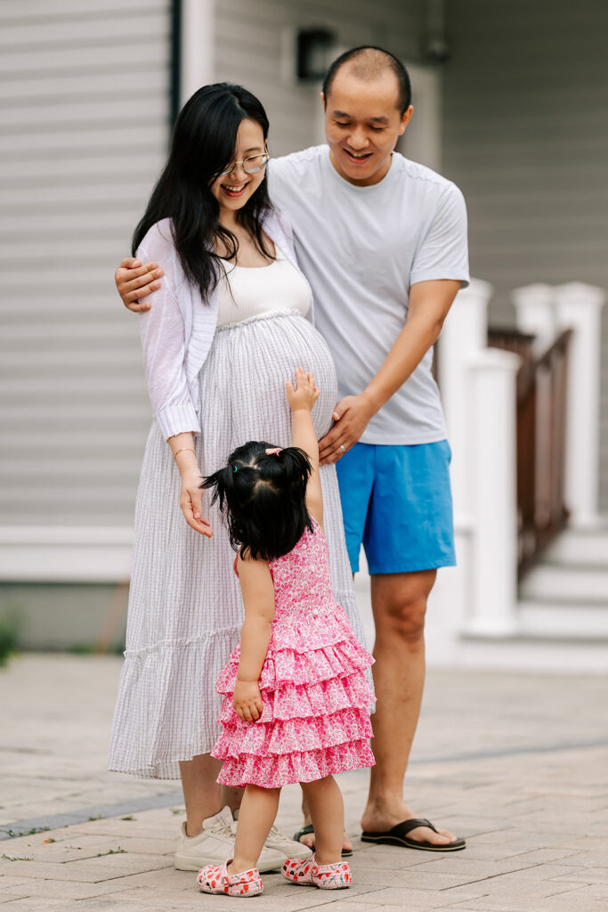 A toddler girl in a pink dress reaches up to touch her mothers bump with both parents looking down on her, on their way to look for baby shower venues in cambridge, MA.